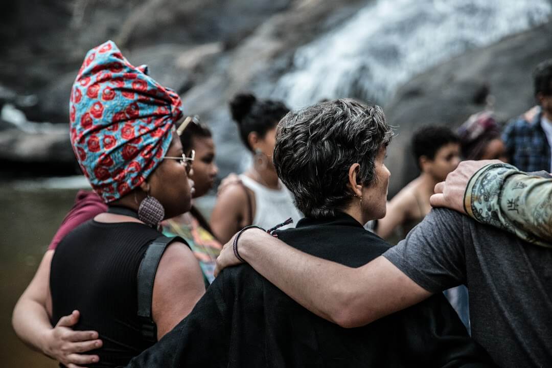 Fotografia mostra um grupo de pessoas que está reunido em roda, de braços dados, em um momento de conexão e acolhimento à beira de uma cachoeira. A imagem é vista de trás e mostra a diversidade do grupo, com destaque para uma mulher negra usando turbante colorido e brincos grandes e outra pessoa com cabelo grisalho curto. A imagem é do grupo do projeto do Movimento dos Ameaçados por Barragens apoiado pelo Programa de Pequenos Projetos da FLD.