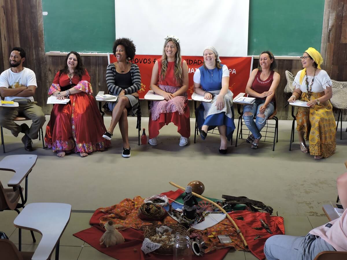 Fotografia de um grupo diverso de oito pessoas está sentado em uma roda, sorrindo e interagindo em um encontro do Comitê dos Povos e Comunidades Tradicionais do Pampa. Ao fundo, há um quadro verde, uma tela branca e um banner do Comitê. Várias pessoas usam roupas tradicionais, incluindo vestidos coloridos, saias estampadas, turbantes e adereços culturais. À frente do grupo, sobre o chão, está um centro com panos coloridos, instrumentos, objetos simbólicos, bonecas e elementos da espiritualidade popular. A cena retrata a importância do trabalho em redes para transformação social.