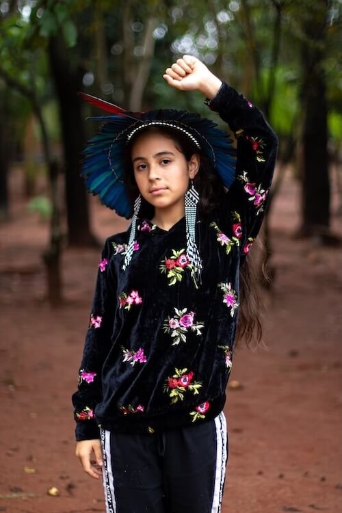 Fotografia de uma menina indígena em pé em uma área de terra batida cercada por árvores. Ela usa um cocar tradicional indígena feito com penas azuis, além de brincos longos de miçangas brancas e pretas. Veste um moletom preto com estampa floral colorida e calça esportiva com listras laterais. A menina olha diretamente para a câmera com expressão firme e levanta o braço direito, com a mão cerrada, em um gesto de afirmação de sua luta por direitos. No canto superior esquerdo, em letras brancas, está escrito: “PROGRAMA COMIN DE defesa de direitos”. A imagem destaca a força e o protagonismo das juventudes indígenas na luta por seus direitos.